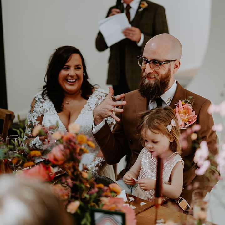 Bride & groom sit with their daughter during industrial styled wedding reception complete with orange fabric table runners and wooden banquet tables
