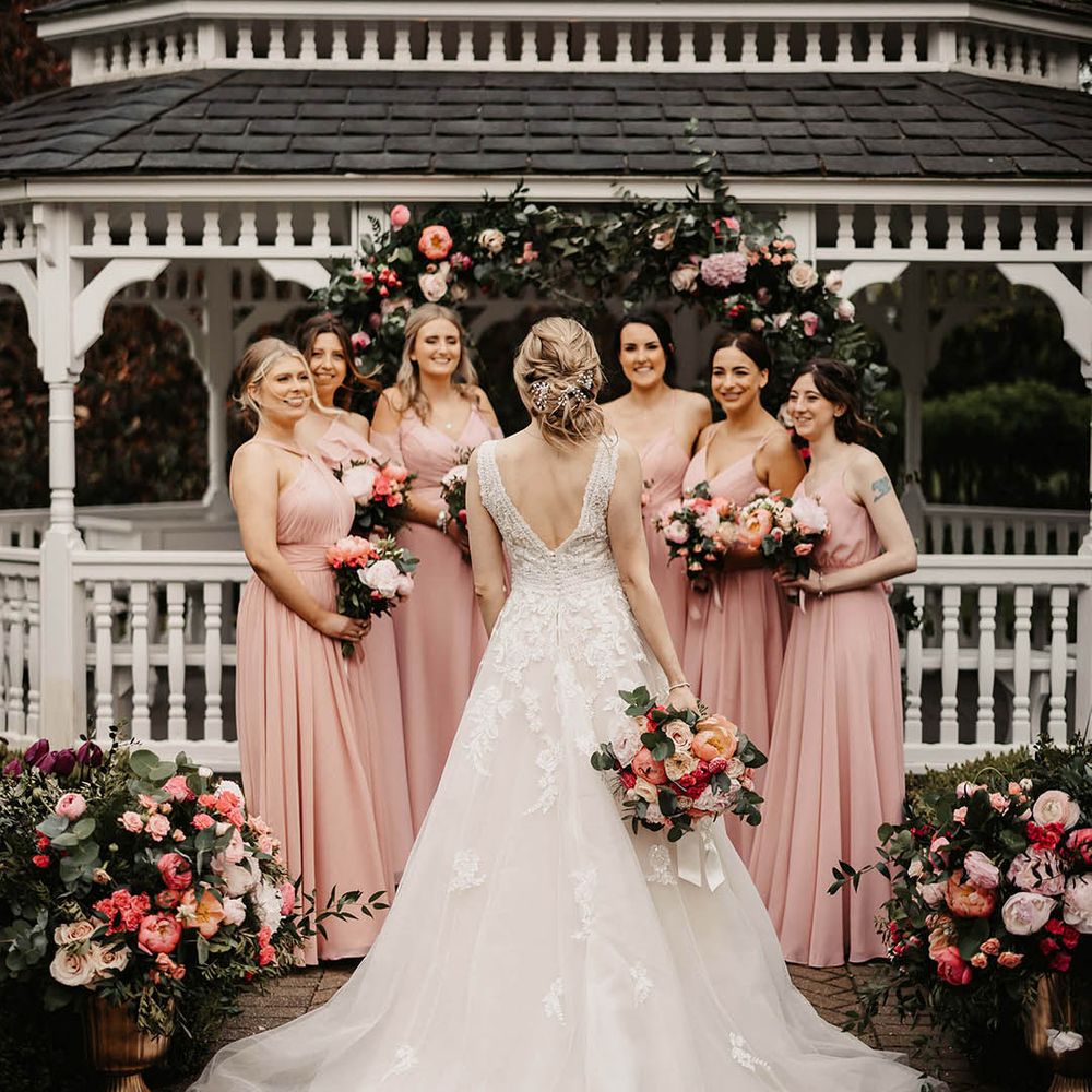 Bridesmaids in pink dresses in front of a pink flower arch holding pink bouquets with the bride showing off her elegant updo 