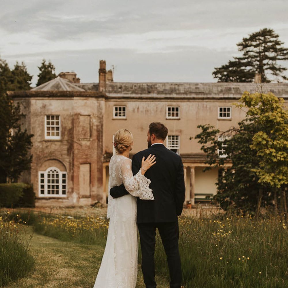 Bride in lace sleeve wedding dress and groom in dark suit embracing on the grounds of Pennard House, one of the best UK country house wedding venues