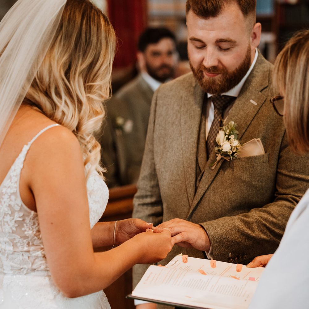 Groom in a grey suit and bride in a lace wedding dress put on their wedding ring for the church ceremony 