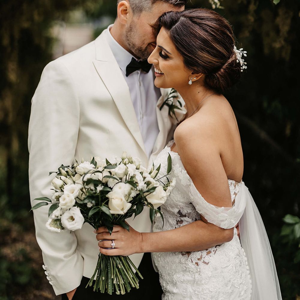 The groom in a white suit jacket embraces the bride in an off the shoulder flower wedding dress holding a white flower bouquet 