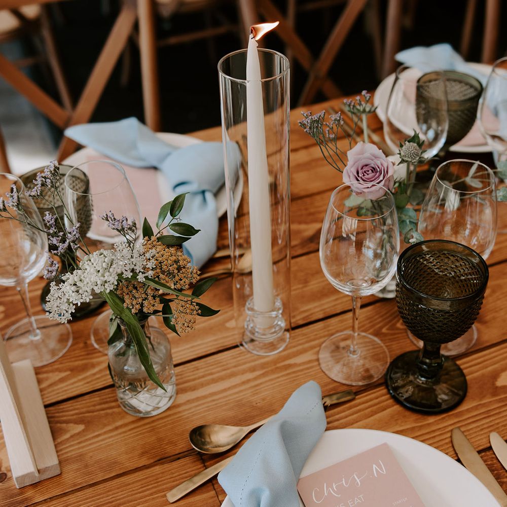 White taper candle in glass vase with white plates, blue napkins and pink wedding menus and gold cutlery