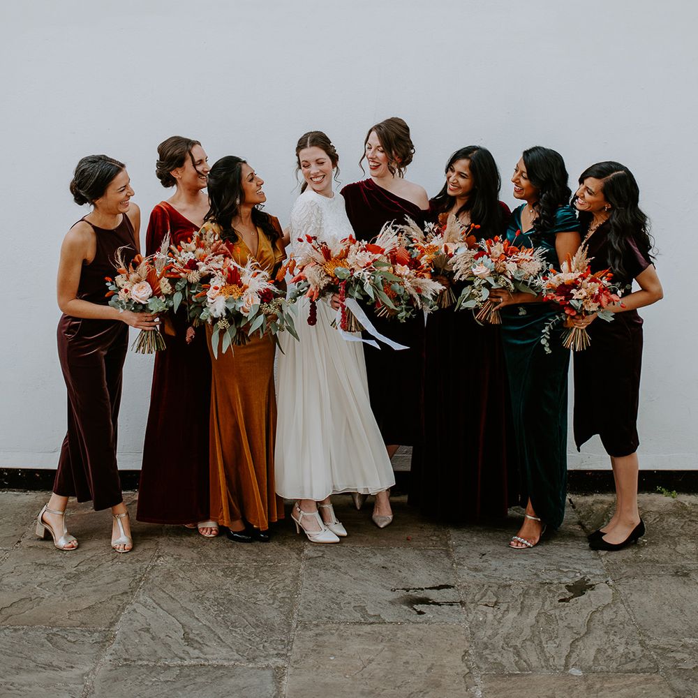 Bride in long sleeve lace vintage wedding dress and closed toe satin heels standing with bridesmaids in mismatched velvet bridesmaid dresses all holding large autumnal bouquets with orange, black, green and yellow roses, peonies, foliage and pampas grass