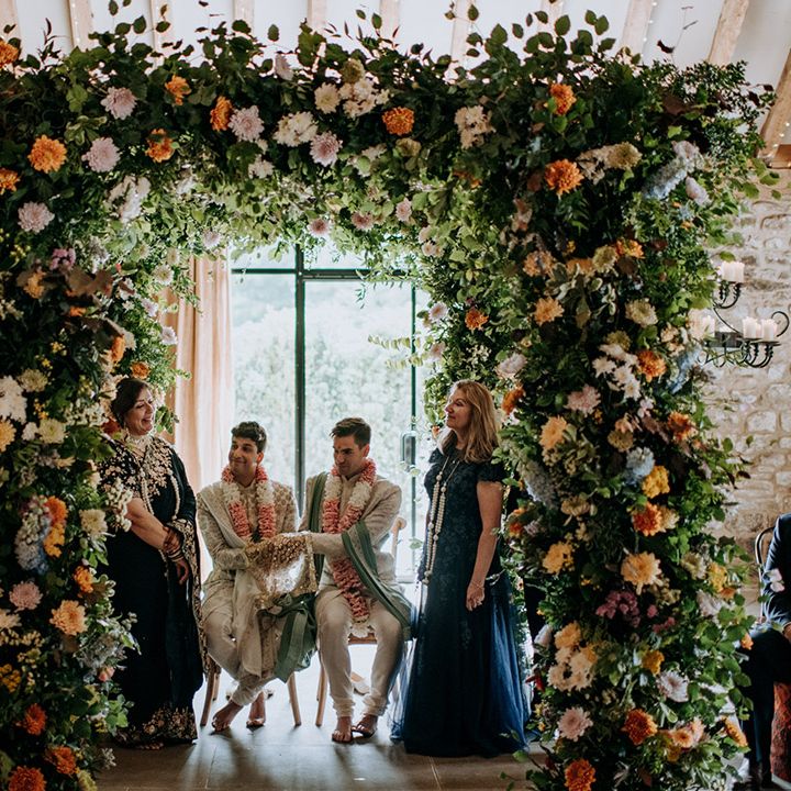Grooms sit between colourful floral arch surrounded by family & friends during wedding ceremony 
