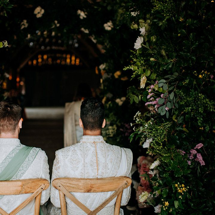 Grooms in matching sherwani's sit together on wooden chairs in the middle of floral arch