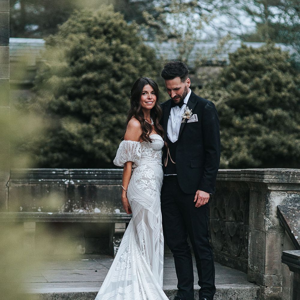 Bride with brown hair in a half up half down style wearing a strapless fitted boho wedding dress standing with the groom in black tie 