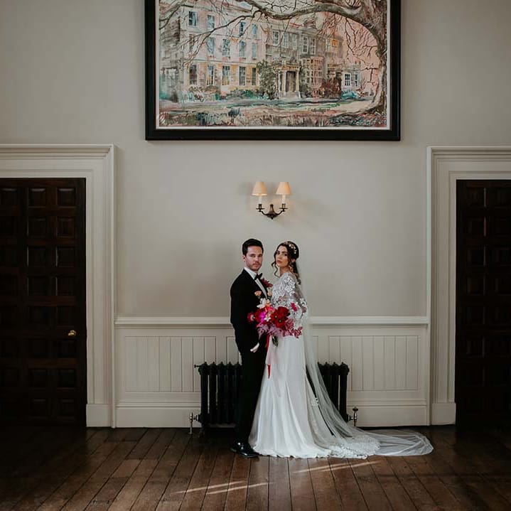 Bride in two piece bridal separates with puff sleeve sheer crop top with 3d appliqué flowers and full bodied silk skirt holding large viva magenta floral bouquet and groom in black tuxedo with magenta amaryllis and orchid boutonniere embracing at Elmore Court wedding venue