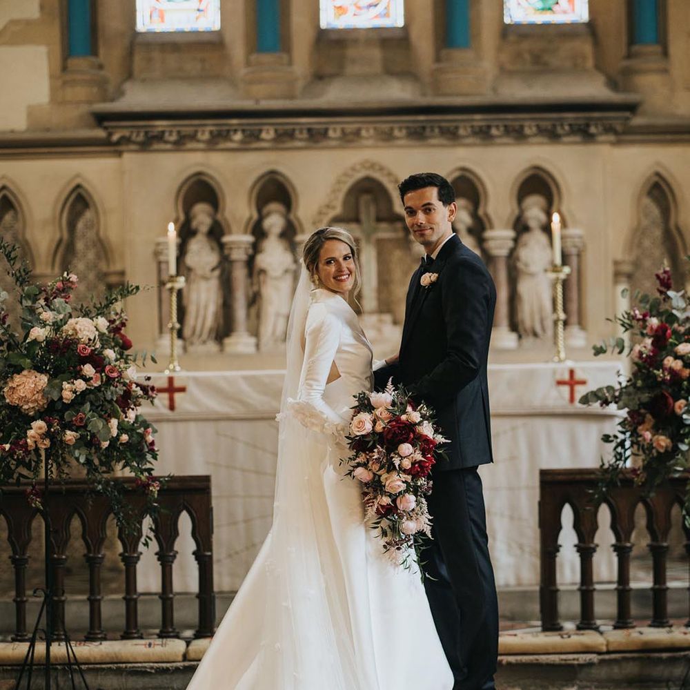 Bride in long sleeve wedding dress with patterned 3D veil standing with the groom in a black tuxedo aa the altar of the church