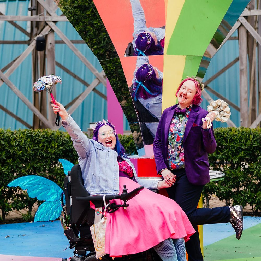 Bride in wheelchair with blue fairy wings wearing personalised denim jacket, pink wedding dress and blue fishnet tights holding hands with bride in purple velvet blazer and black suit trousers with purple bowtie, pink patterned pocket square and colourful patterned shirt on rainbow floor 