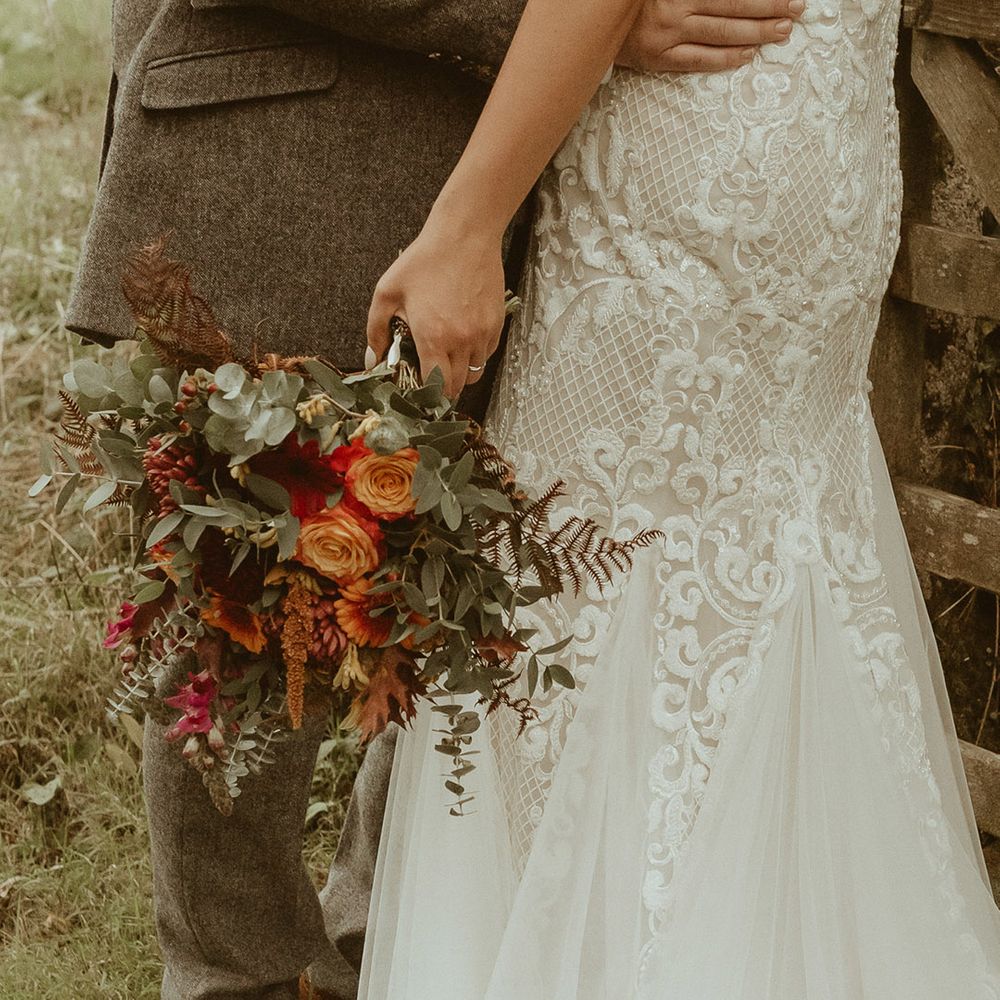Bride in fitted lace off the shoulder boho wedding dress holding orange autumnal bouquet with the groom in a three piece tweed suit 