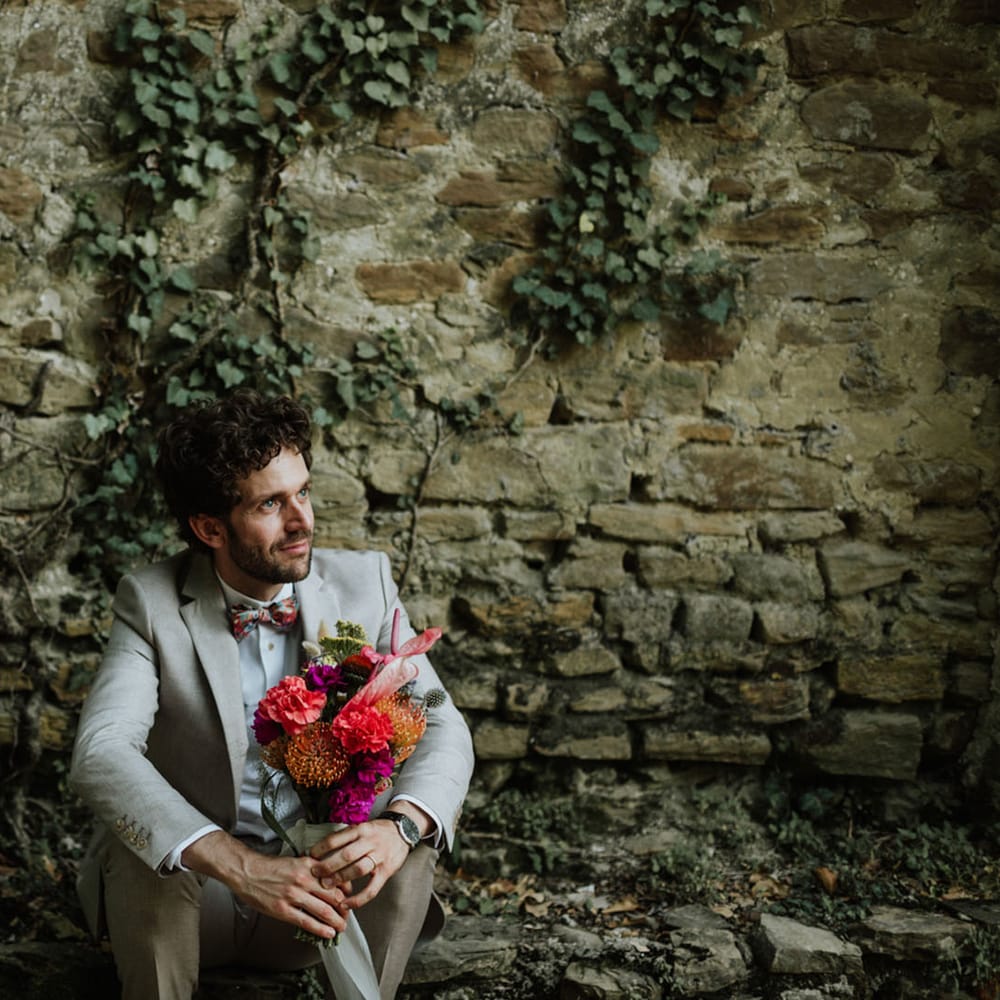 Groom in greige suit and colourful bow tie sitting down with bridal bouquet consisting of red pincushion flowers, coral carnations, magenta peonies and anthuriums