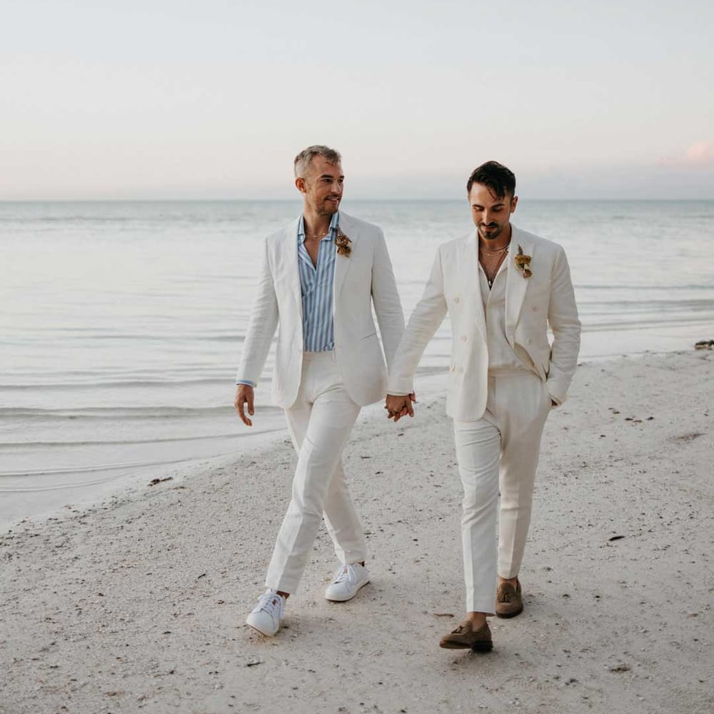 Groom wearing white blazer, light blue and white striped shirt, dusky orange garden rose and dried flower boutonniere holding hands with groom in mens white wedding suit with dusky orange garden rose and dried flower boutonniere, white shirt and silver jewellery 