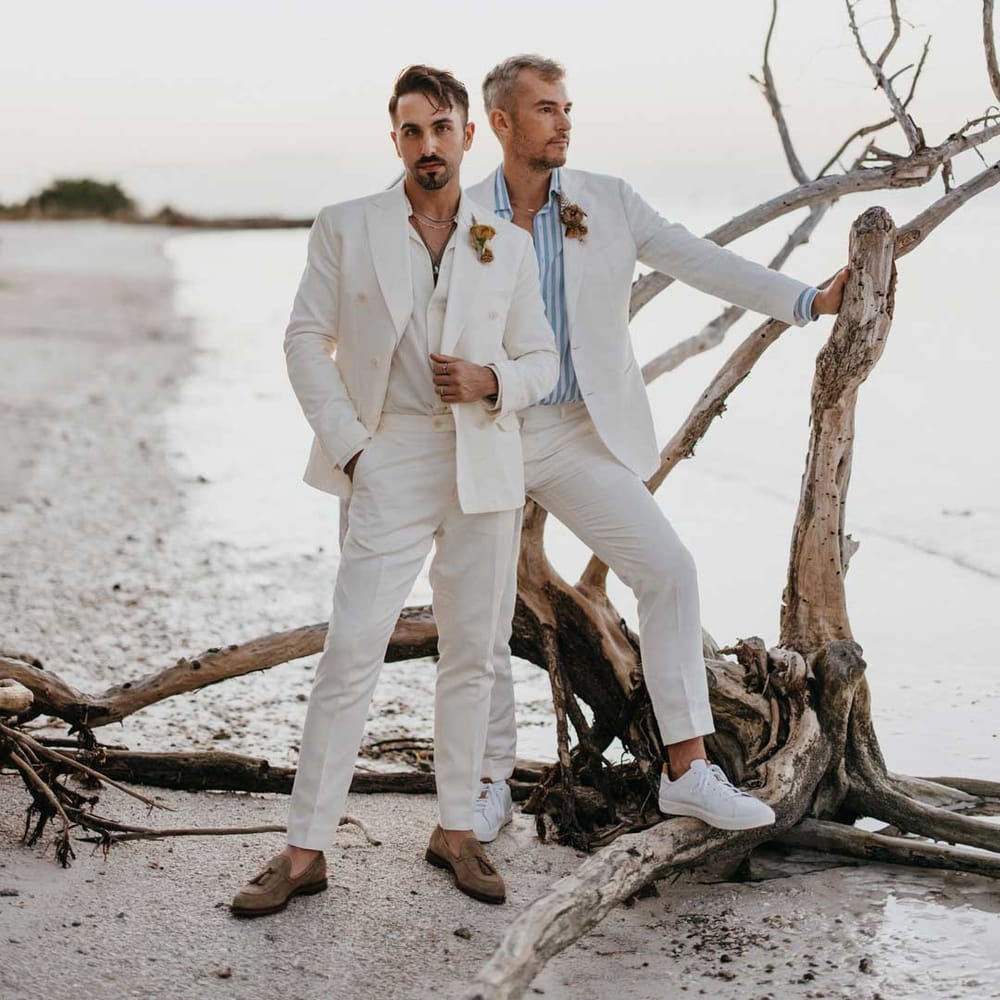 Groom in mens white wedding suit with dusky orange garden rose and dried flower boutonniere, white shirt and silver jewellery posing with groom in white blazer, light blue and white striped shirt, dusky orange garden rose and dried flower boutonniere and gold jewellery posing by some driftwood 
