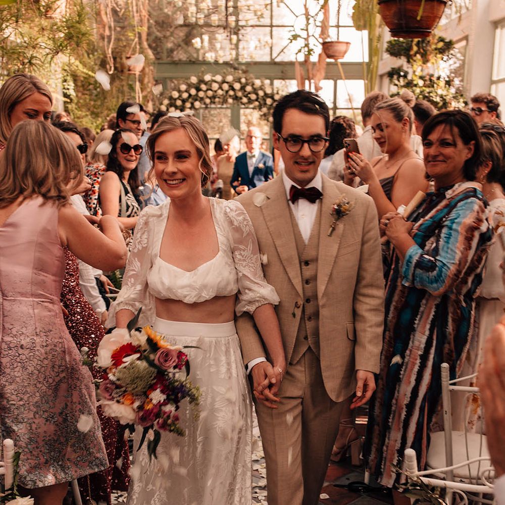 Bride in lace bridal separates walking back down the aisle with the groom in a cream suit as a married couple to white petal confetti 