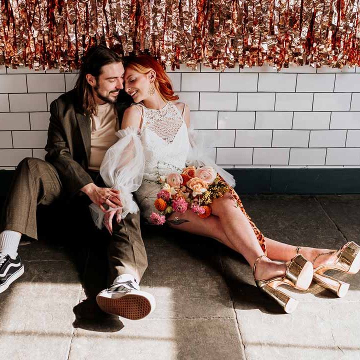 Groom in brown blazer, white shirt, grey suit trousers and vans shoes sitting on the floor of Northern Monk Refectory Leeds with bride in 70s inspired boatneck short lace wedding dress with detachable tulle puff sleeves and platform gold wedding heels 
