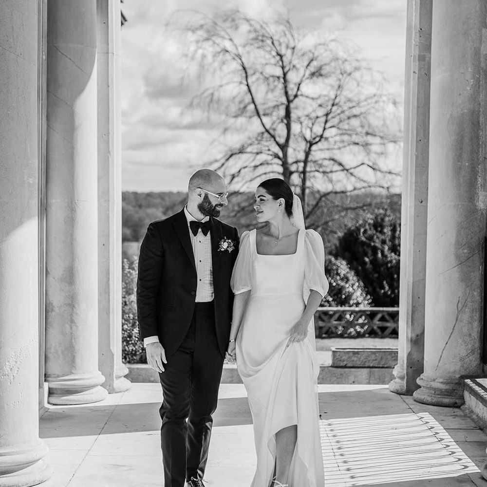 Bride in short sheer puff sleeve wedding dress with square neck and wrist loop with sheer pearl embellished bridal veil walking with groom in classic black tuxedo with bowtie and wildflower boutonniere at Buxted Park