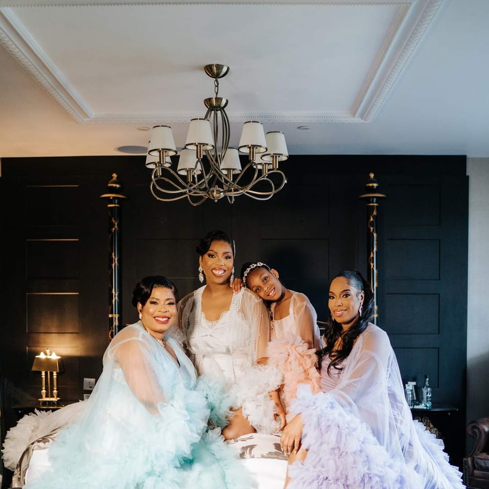 Bride in white sheer tulle and pearl bridal getting ready robe with bridesmaids and flower girl in mismatched pastel sheer tulle and pearl bridal getting ready robes sitting on bed at Syon Park wedding venue