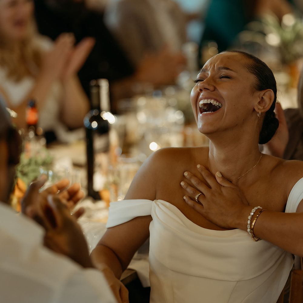 bride-laughing-in-off-the-shoulder-wedding-dress-listening-to-wedding-speeches