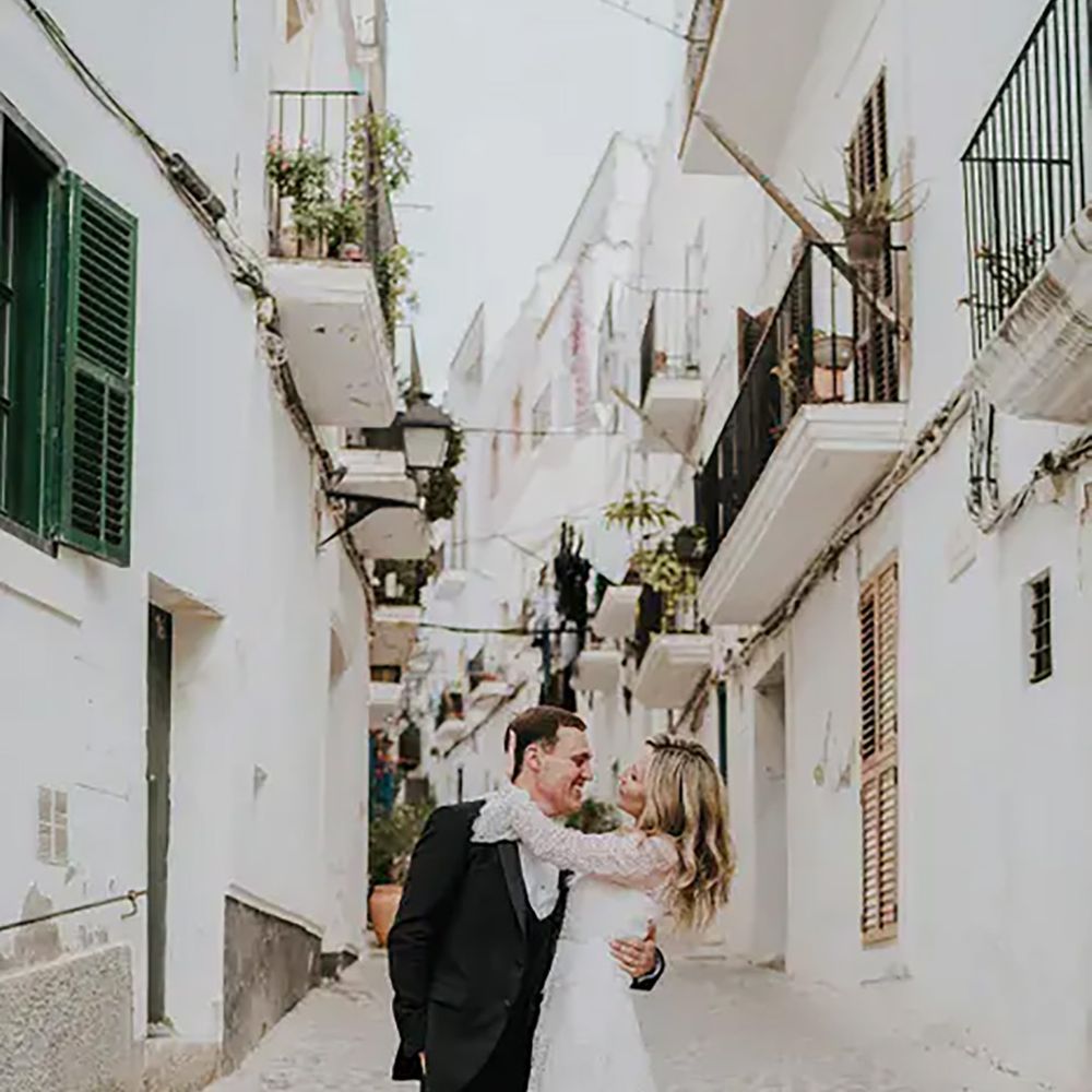 bride-in-a-boho-lace-wedding-dress-with-long-sleeves-cupping-her-grooms-head-in-the-side-streets-of-ibiza-with-white-buildings-balconies-and-shutters