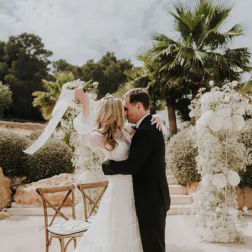 bride-in-a-long-bell-sleeve-lace-wedding-dress-waving-her-white-wedding-bouquet-tied-with-ribbon-in-the-air-as-she-kisses-her-groom-in-a-tuxedo-at-pure-house-ibiza-wedding