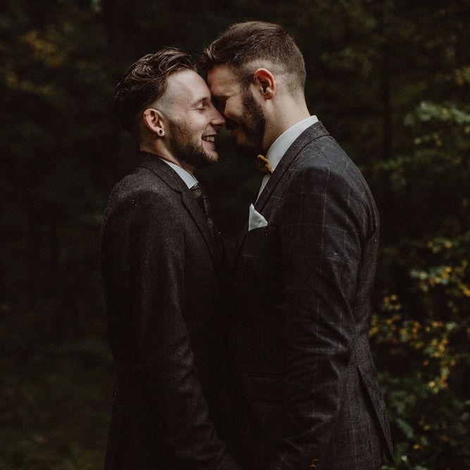 Two grooms stand with foreheads together smiling in dark wedding photo
