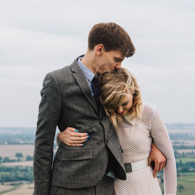 Bride and Groom stand side by side with arms around each other, groom kisses bride on the crown of her head