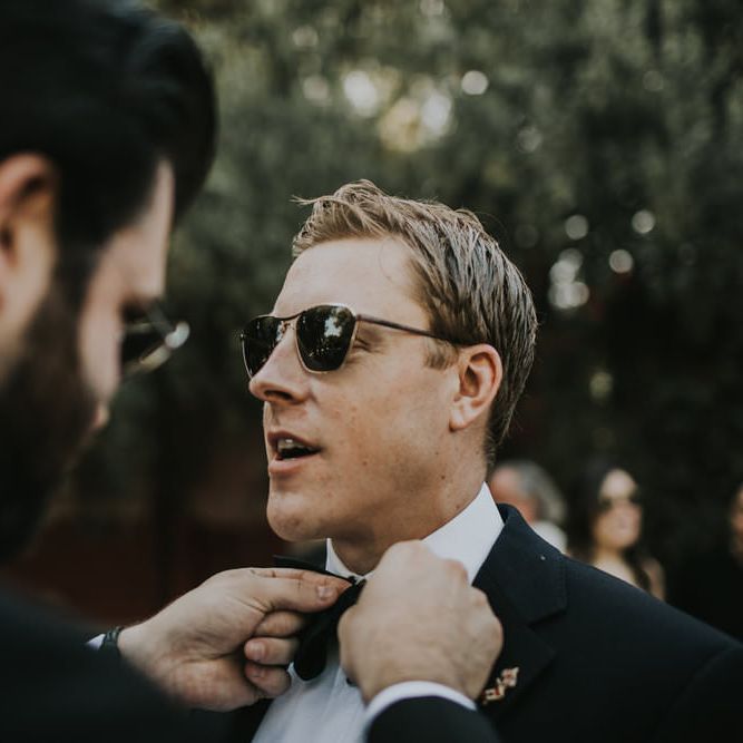 Groomsman with dark beard adjusts the bow tie on a white groom wearing sunglasses