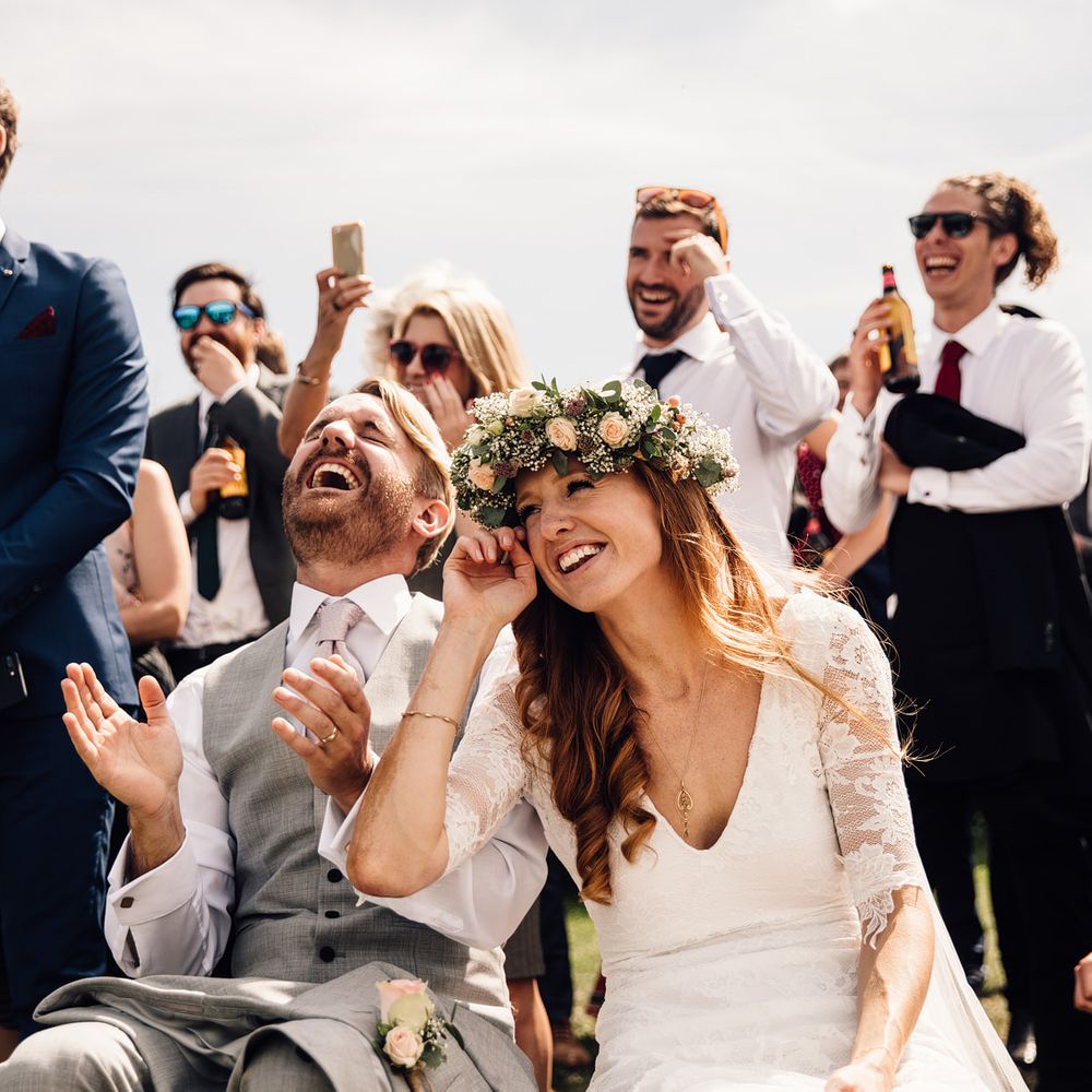 Groom in grey waistcoat laughs and claps hands with bride wearing flower crown next to him and guests smiling behind - Documentary Style Wedding Photography