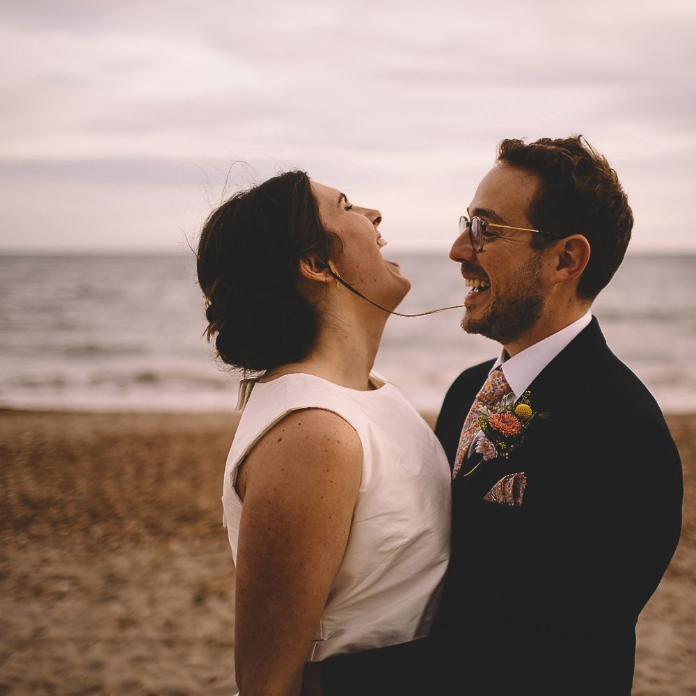 Bride in Homemade Wedding Dress with Bow Back and Groom in Dress2Kill Suit Laughing on the Beach