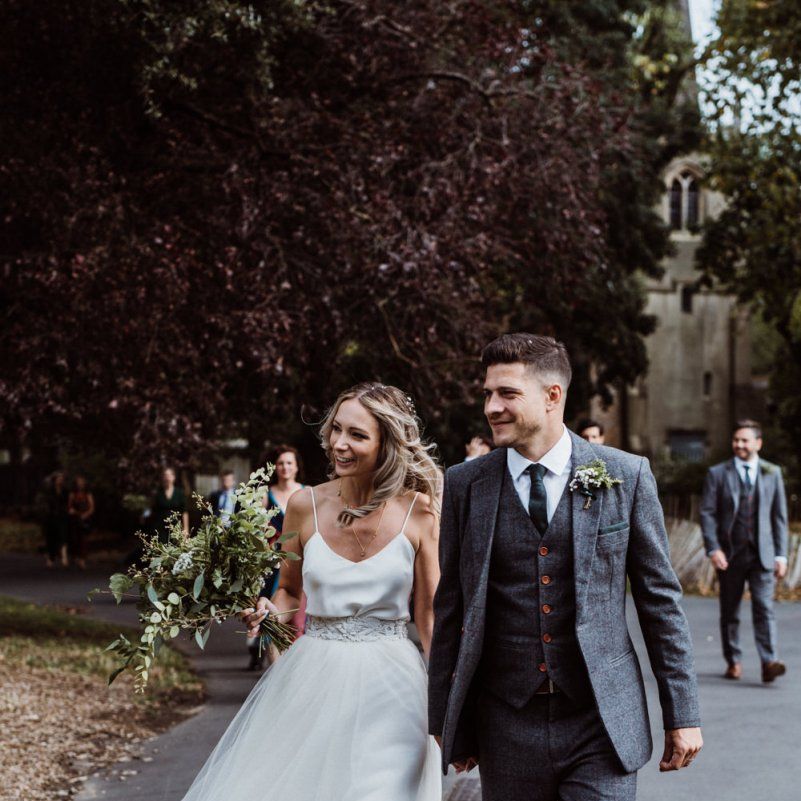 Bride and groom walk together to their pub reception wearing two piece gown and foliage bouquet