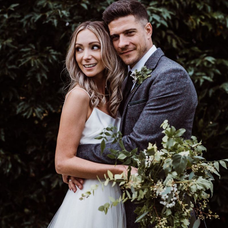 Bride and groom embrace whilst clutching foliage bouquet