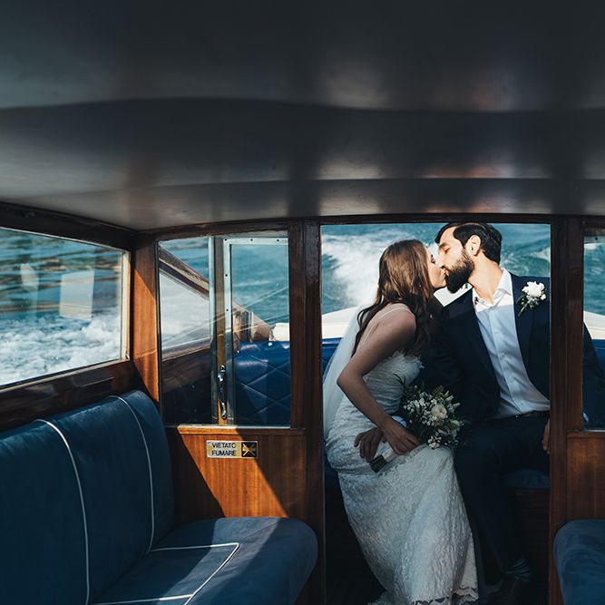 Bride in Grace Loves Lace Mia Wedding Dress and Groom in Taliare Navy Suit Kissing on Boat Ride Over Lake Como