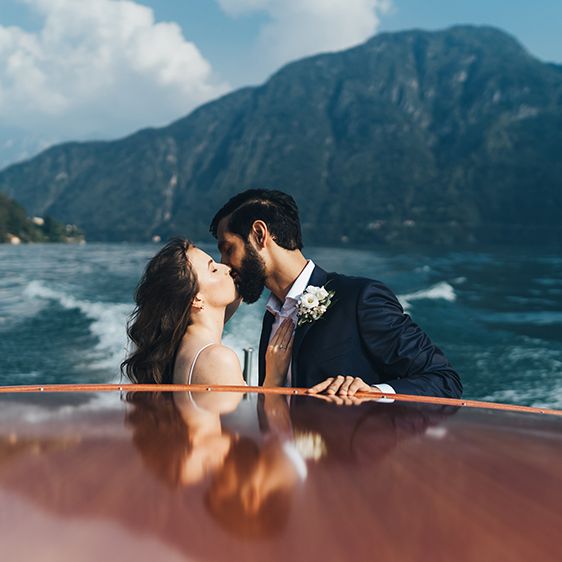 Bride in Grace Loves Lace Mia Wedding Dress and Groom in Taliare Navy Suit Kissing on Boat Ride Over Lake Como