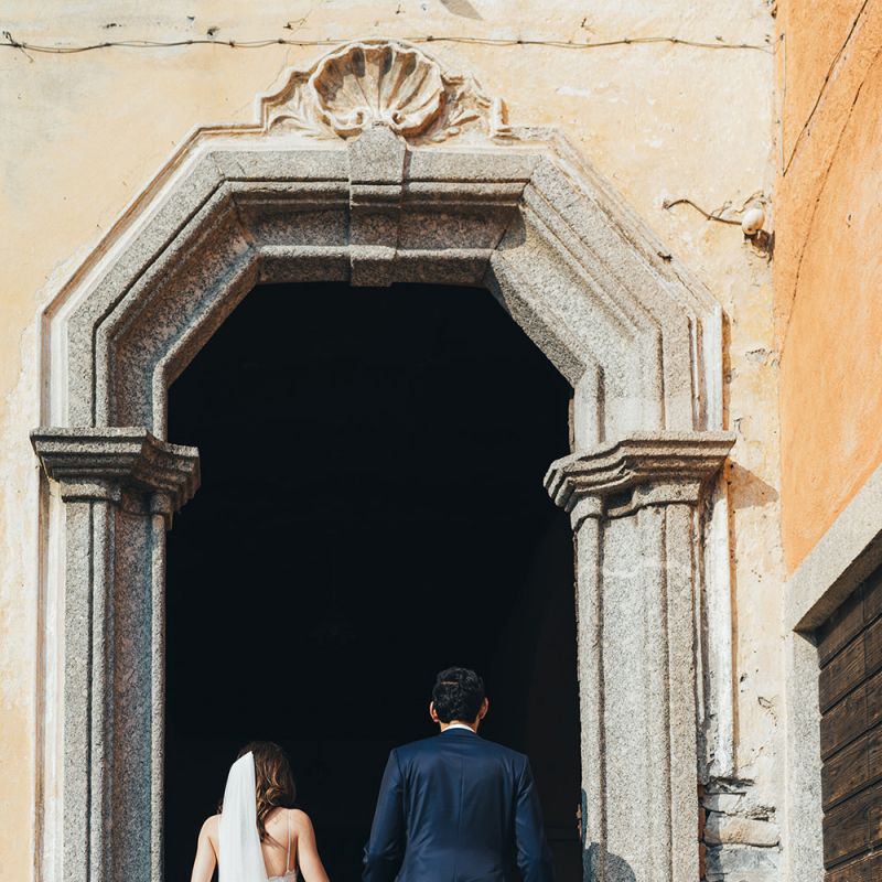 Bride in Grace Loves Lace Mia Wedding Dress and Groom in Taliare Navy Suit Entering Wedding Ceremony Venue