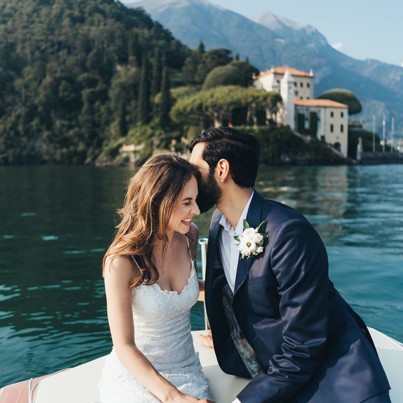 Laughing Bride in Grace Loves Lace Mia Wedding Dress and Groom in Taliare Navy Suit Enjoying a Boat Ride Over Lake Como