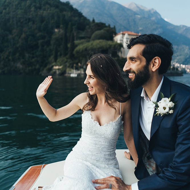 Waving Bride in Grace Loves Lace Mia Wedding Dress and Groom in Taliare Navy Suit Enjoying a Boat Ride Over Lake Como