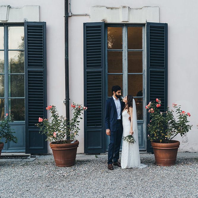 Bride in Grace Loves Lace Mia Wedding Dress and Groom in Taliare Navy Suit Standing in Front of giant Window Shutters