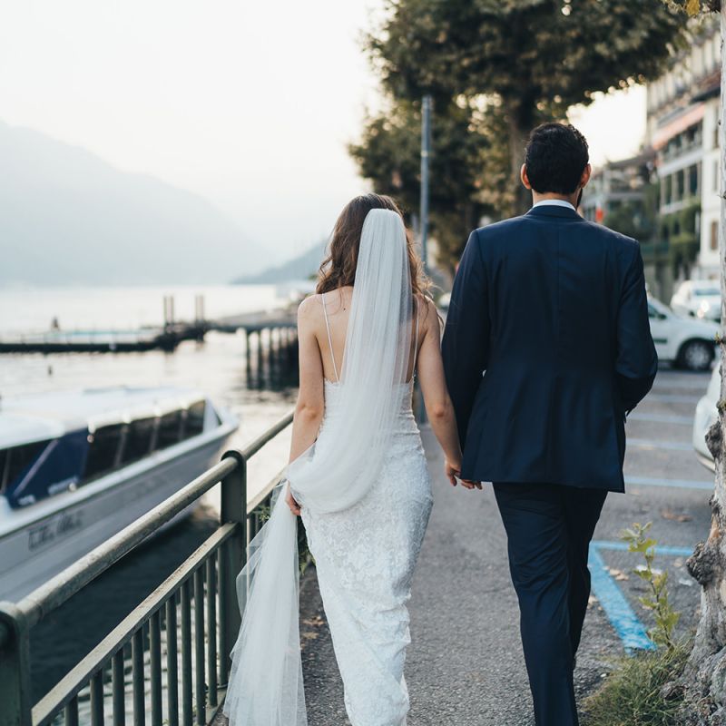 Bride in Grace Loves Lace Mia Wedding Dress and Groom in Taliare Navy Suit Walking Around Lake Como