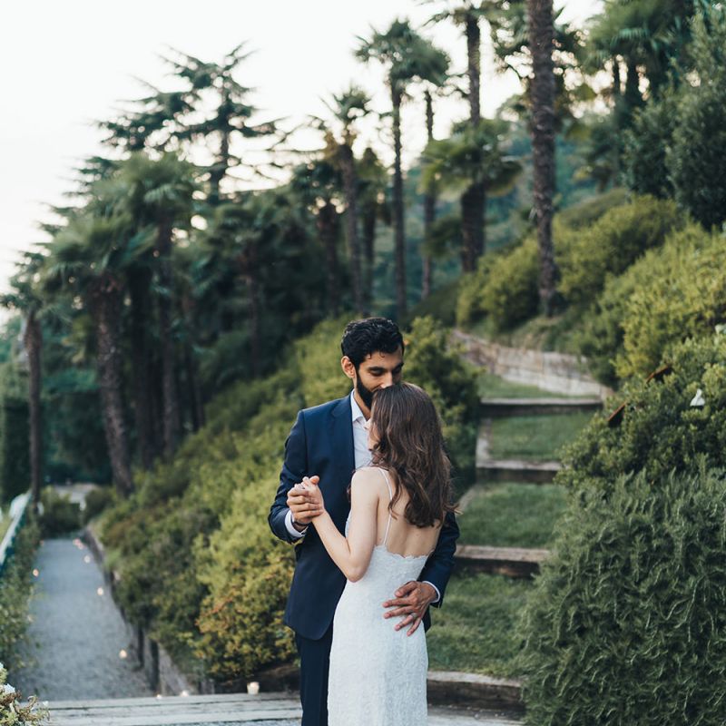 Bride in Grace Loves Lace Mia Wedding Dress and Groom in Taliare Navy Suit Embracing in Gardens