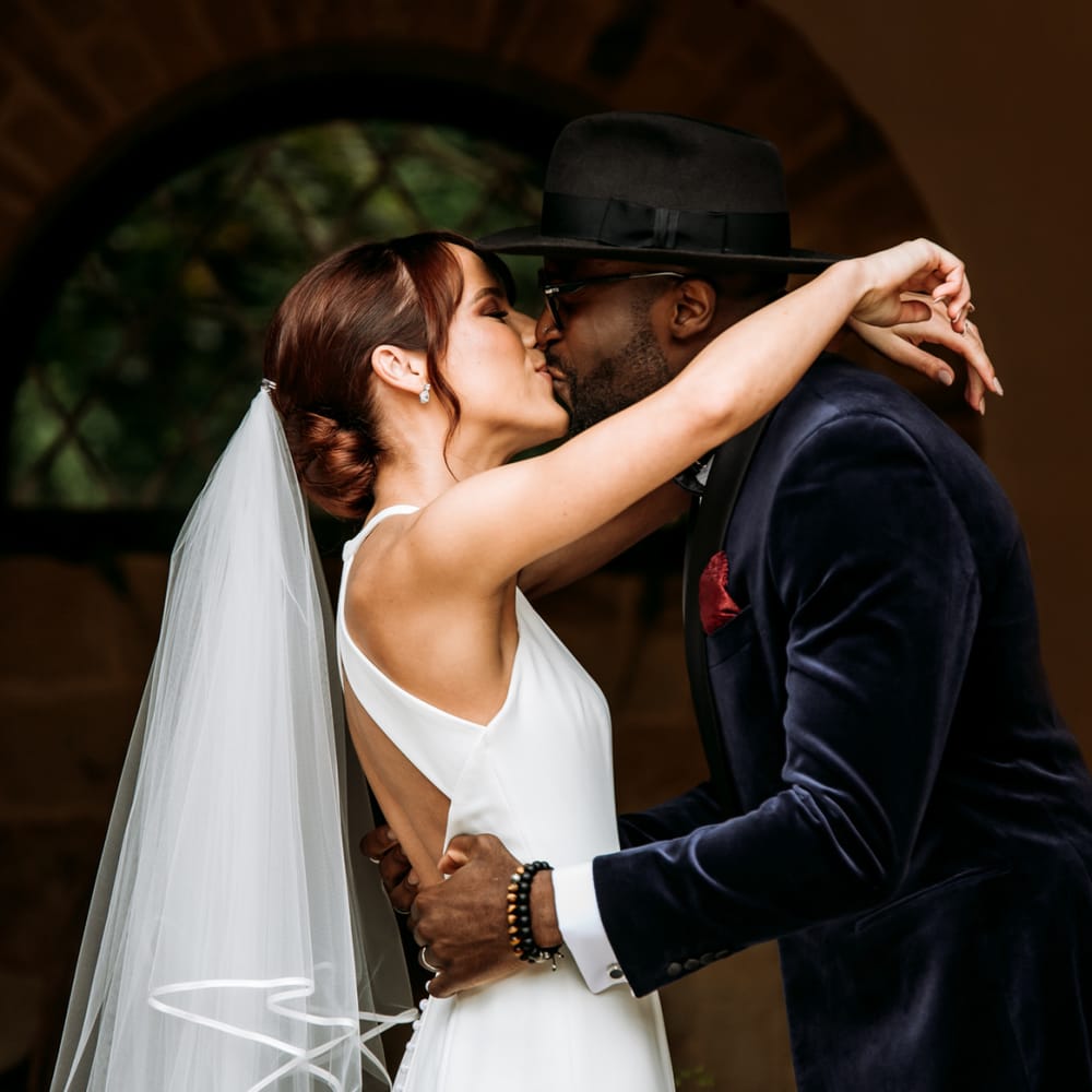 Groom in velvet dinner jacket and fedora kissing his bride at The Ravenswood wedding