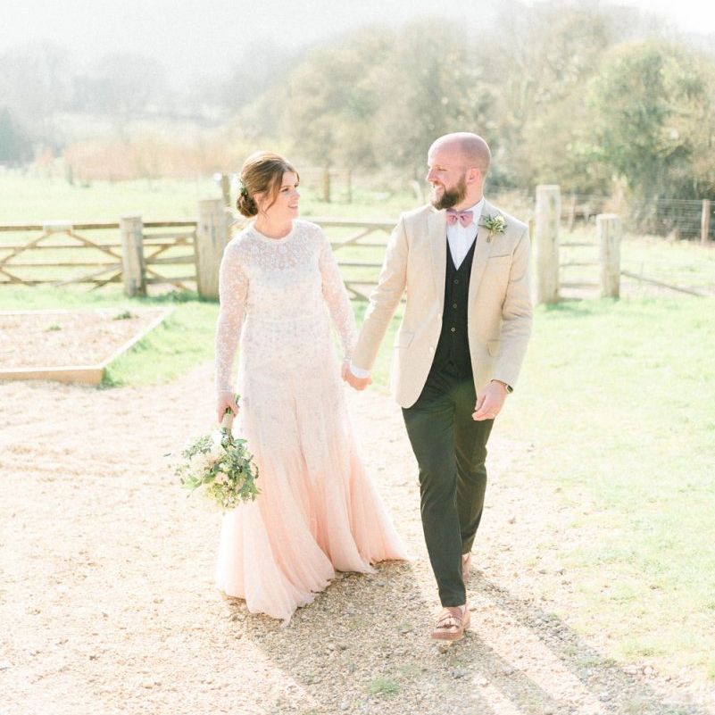 Bride in Needle &amp; Thread Wedding Dress and Groom in Beige Blazer and Pink Bow Tie Holding Hands