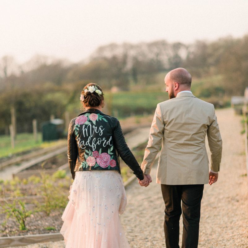 Groom in Black Trousers and Beige Blazer and Bride in Hand Painted Leather Jacket Hand in Hand