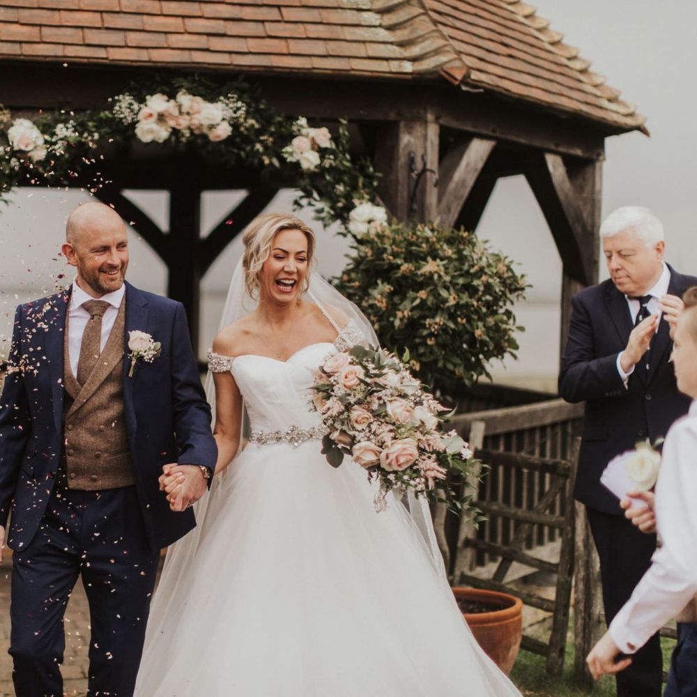 Bride and groom at outdoor ceremony with pink rose bouquet and off the shoulder bridal dress with embellished belt detail