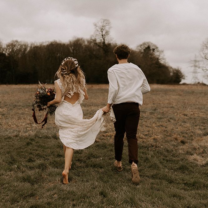 Boho Bride in Lace Wedding Dress and Groom in Casual Shirt and Trousers Running Through the Field