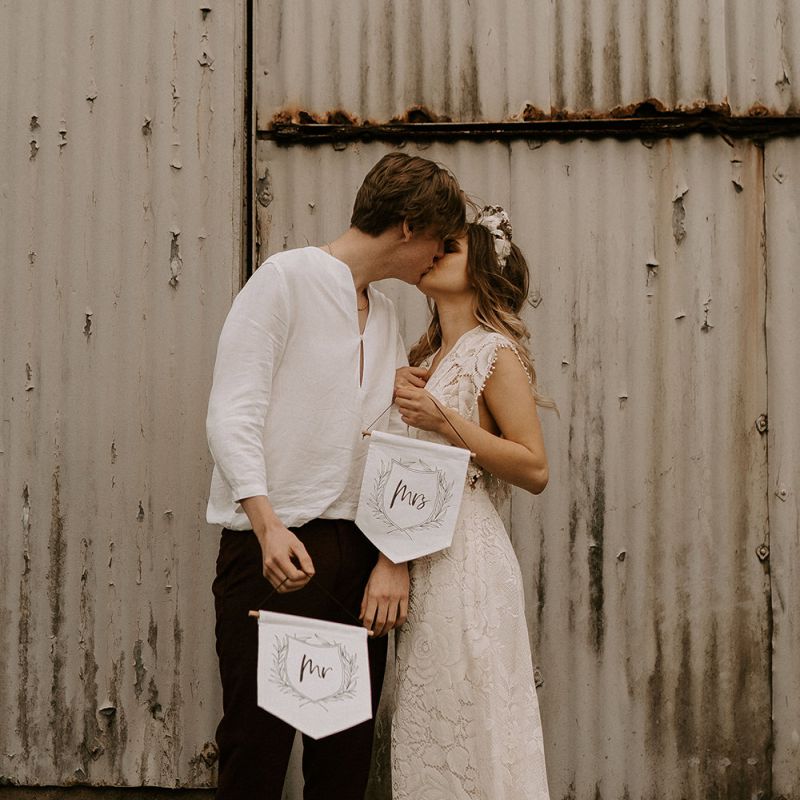 Boho Bride in Lace Wedding Dress and Groom in Casual Trousers and Shirt Holding Up Mr &amp; Mrs Banners