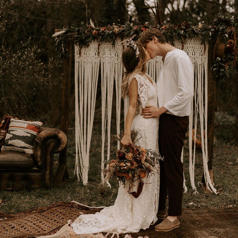 Boho Bride in Lace Wedding Dress Kissing Her Groom in Casual Shirt and Trousers at the Altar of Macrame Backdrop and Persian Rug Floor