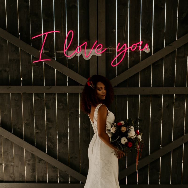 Black Bride with Afro Hair in Lace Wedding Dress Holding a Red and White Wedding Bouquet Under a Neon Wedding sign