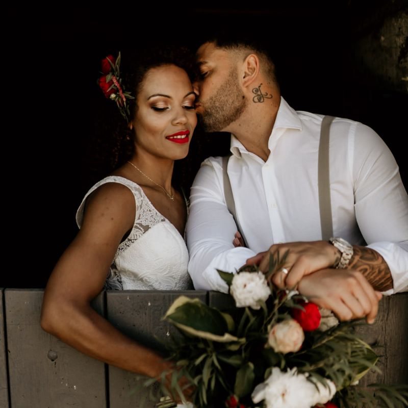 Black Groom in White Shirt and Braces Kissing Bride with Afro Hair in a Lace Wedding Dress