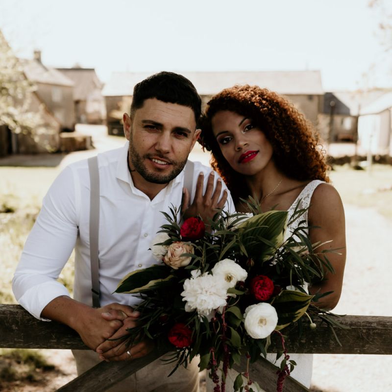 Black Bride in Lace Wedding Dress and Groom in White Shirt and Braces Embracing Next to Country Fence