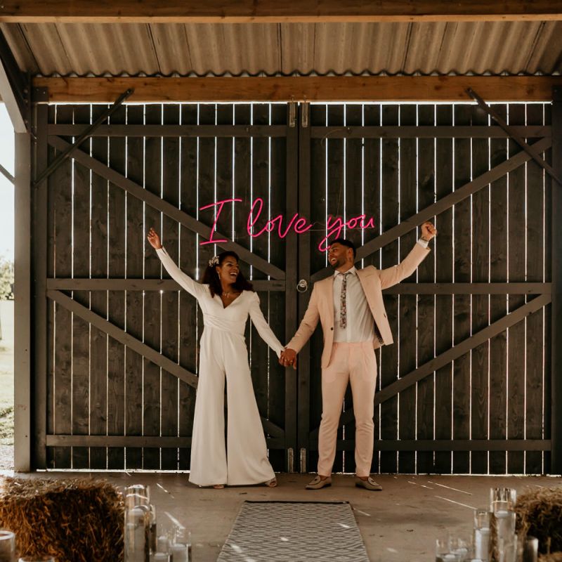 Black Bride with Afro Hair  in White Jumpsuit with Groom in Pink Suit Standing Next to Neon I Love You Sign